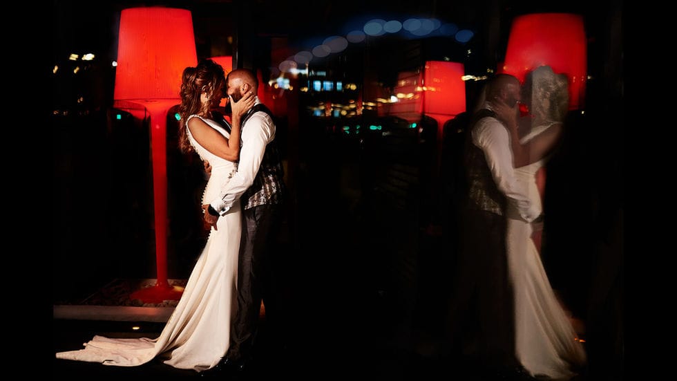 Bonita foto de Postboda, en la que los novios se besan y están delante de un cristal en donde su reflejo marca la simetría por detrás hay unas lámparas rojas que aún marcan más las diagonales y la simetría.