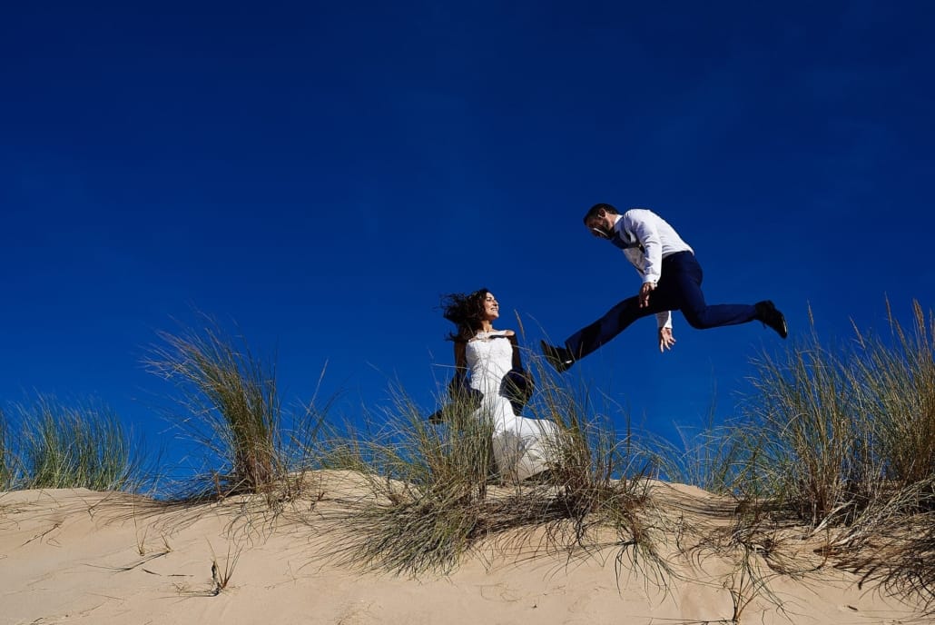 En las dunas de la playa salve de Laredo unos novios el día de su Postboda, posan para nosotros ,la novia mira como el novio salta por encima de la duna y los dos están recortados en un intensísimo cielo azul que contrasta con la arena dorada