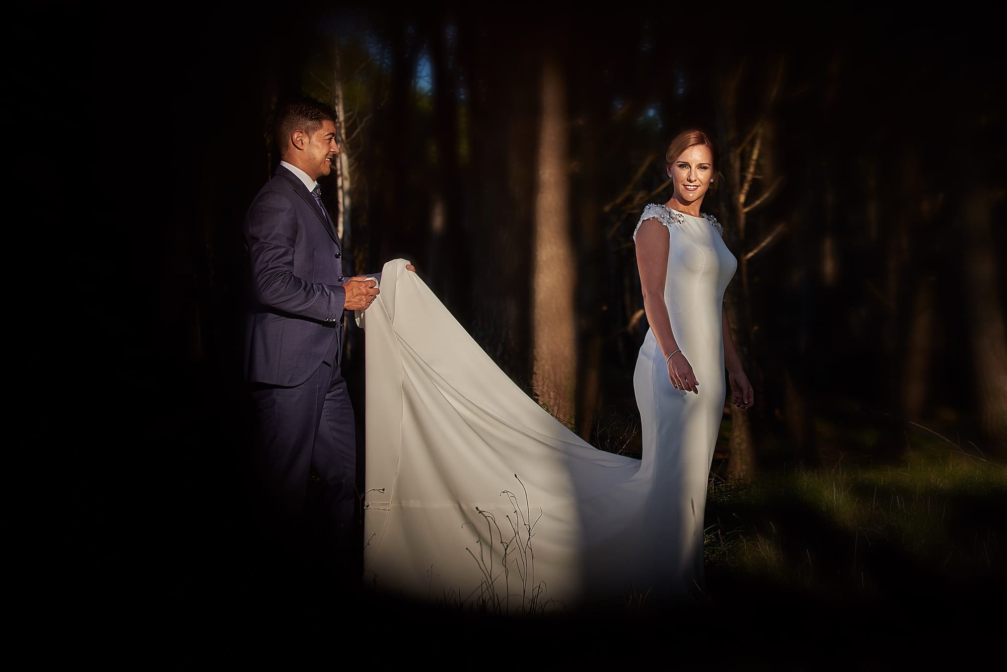 Romántica fotografía en los pinares de Liencres al atardecer, jugando con los luces y las sombras en la que el novio sujeta la cola del vestido de la novia y ella luce una espléndida silueta