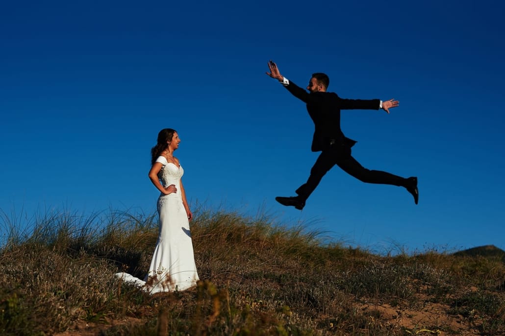 En las dunas de Liencres contra un cielo azul intenso y un ocre de arena, los novios posan en su Postboda ella mira el novio y el novio hace un salto divertido recortándose contra el cielo