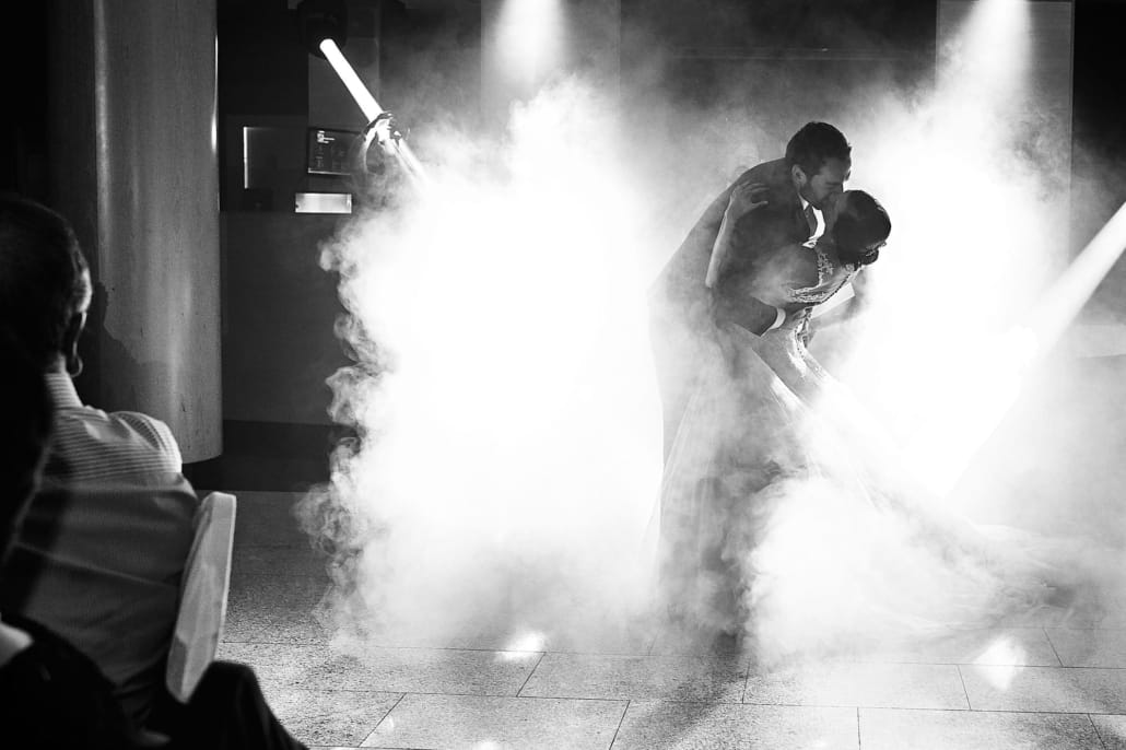 124-JOSE FERREIRO FOTOGRAFIA - FOTOGRAFO DE BODAS EN SANTANDER Fotografía en blanco y negro de la entrada de los novios al comedor, el comedor está a oscuras y solo hay efectos de luces y niebla. La foto muestra a los novios recortándose en la niebla y besandose en las entrada al comedor