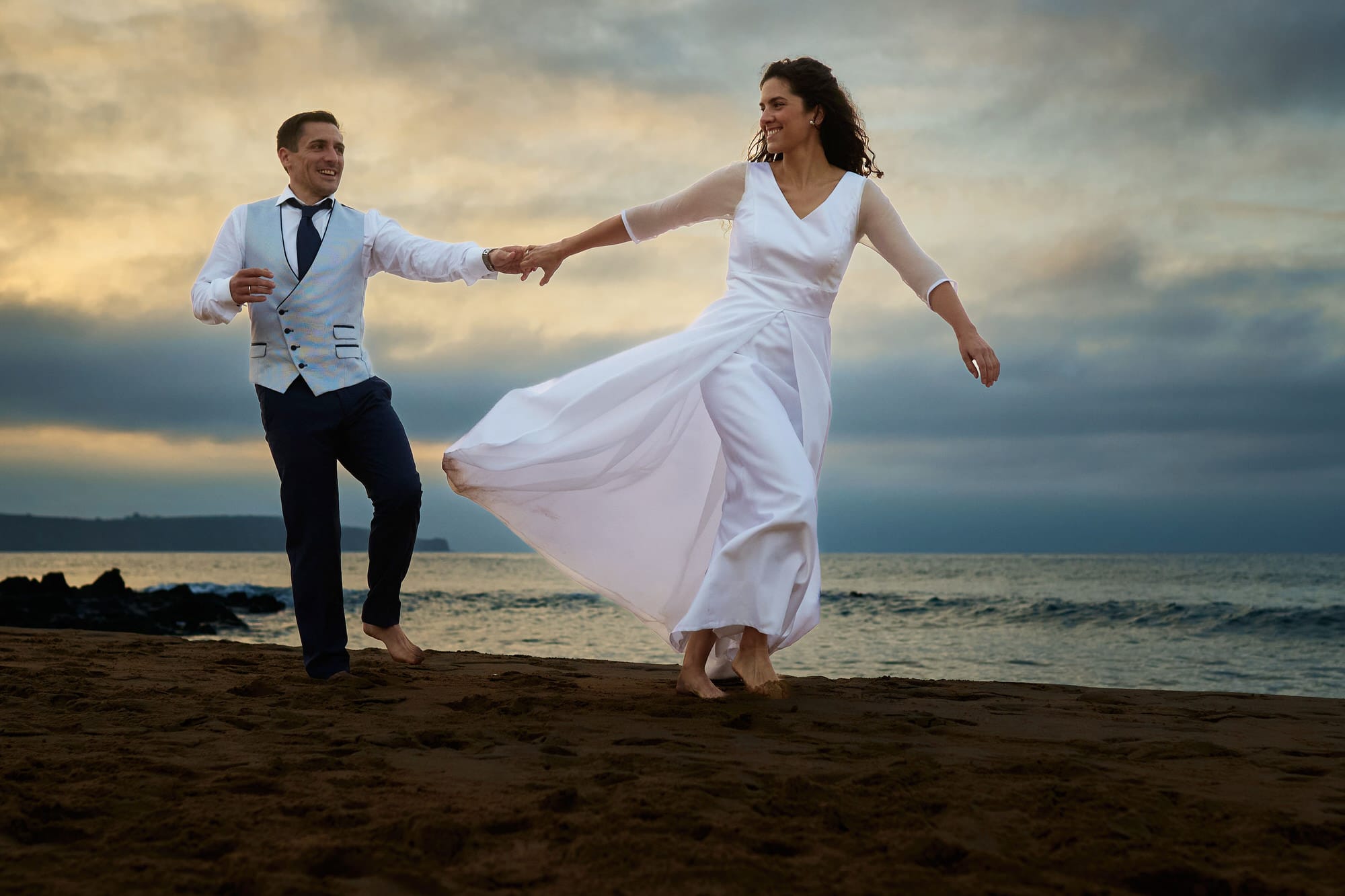 Dos novios corren descalzos vestidos con sus trajes de boda, ella con un vestido blanco por delante de el, en una puesta de sol sobre la arena en la playa de liencres