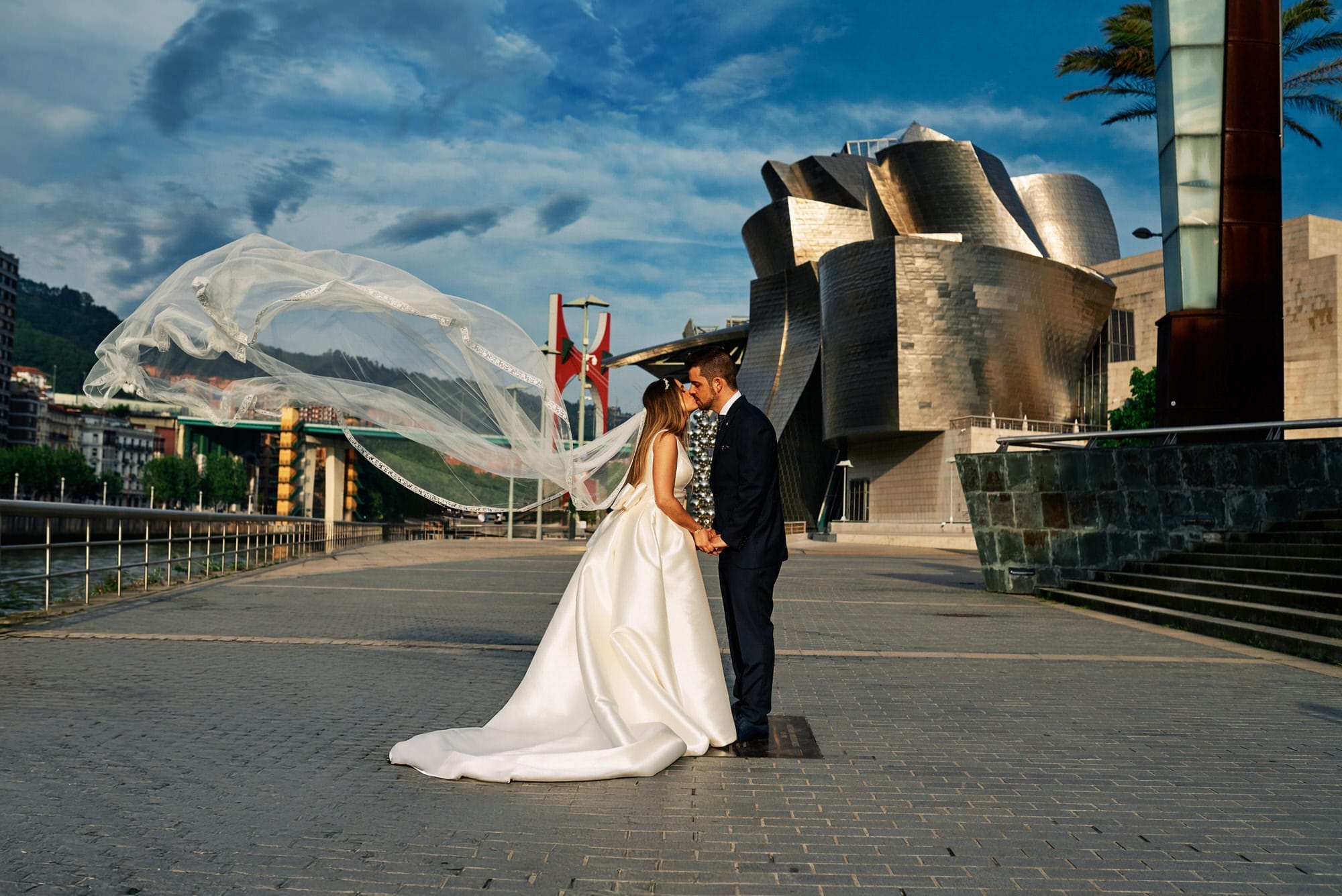 Dos novios besándose vestidos el día de su boda frente al Guggenheim. El viento levanta el velo de la novia.