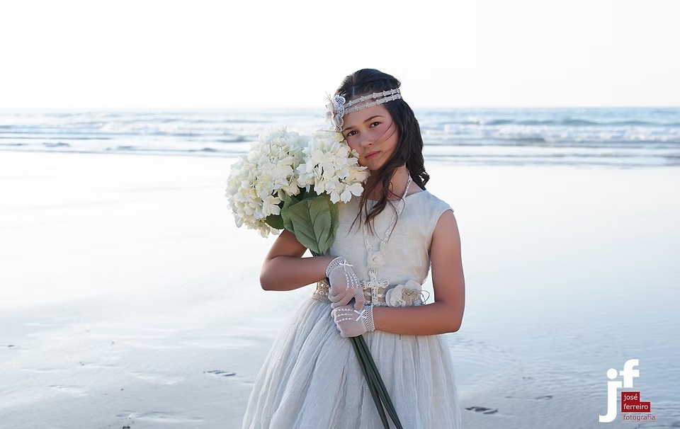 comuniones jose ferreiro 2026-13 Una niña vestida de comunión con un ramo de flores en sus manos, está posando en la playa para nosotros