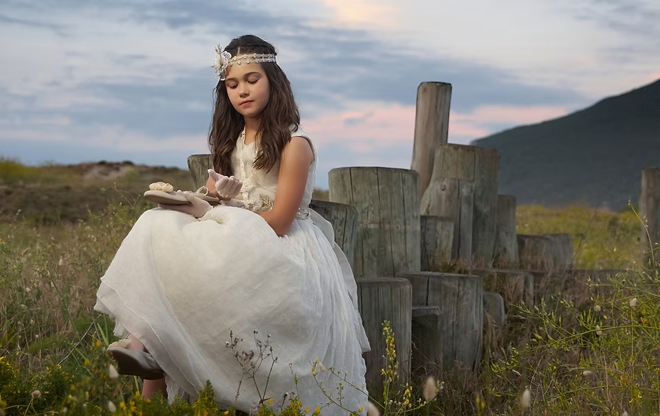 comuniones jose ferreiro 2026-6 Niña vestida de comunión está sentada en unos troncos de madera y mira detenidamente sus zapatos