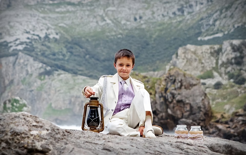 comuniones jose ferreiro 2026-9 Un niño sentado en una roca, con unos acantilados, al fondo está vestido de comunión, sujeta un farol