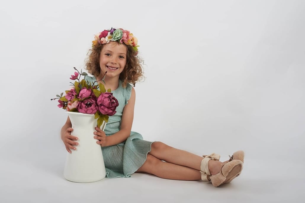 fotografia infantil, la niña no mira , esta sentada, tiene una jarra blanca con peonias y lleva una diadema de flores