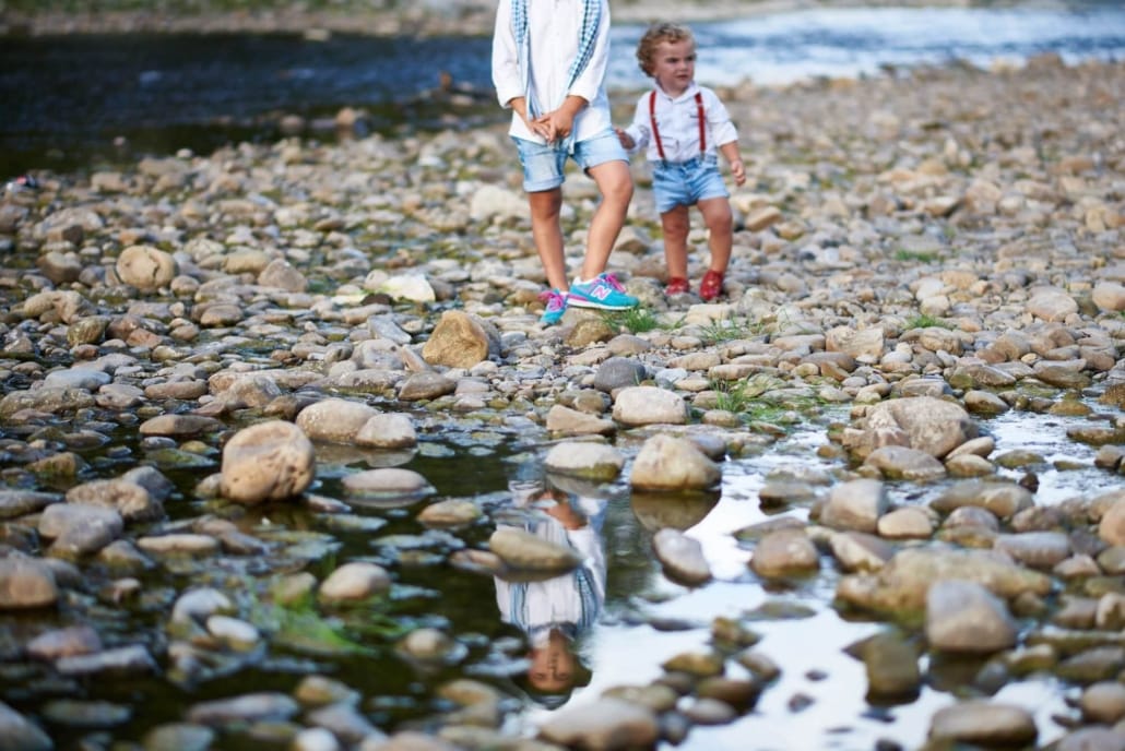 foto de son niños pequeños sobre las piedras del rio, se ve al pqño de la mano del mayor y este se refleja en un charco
