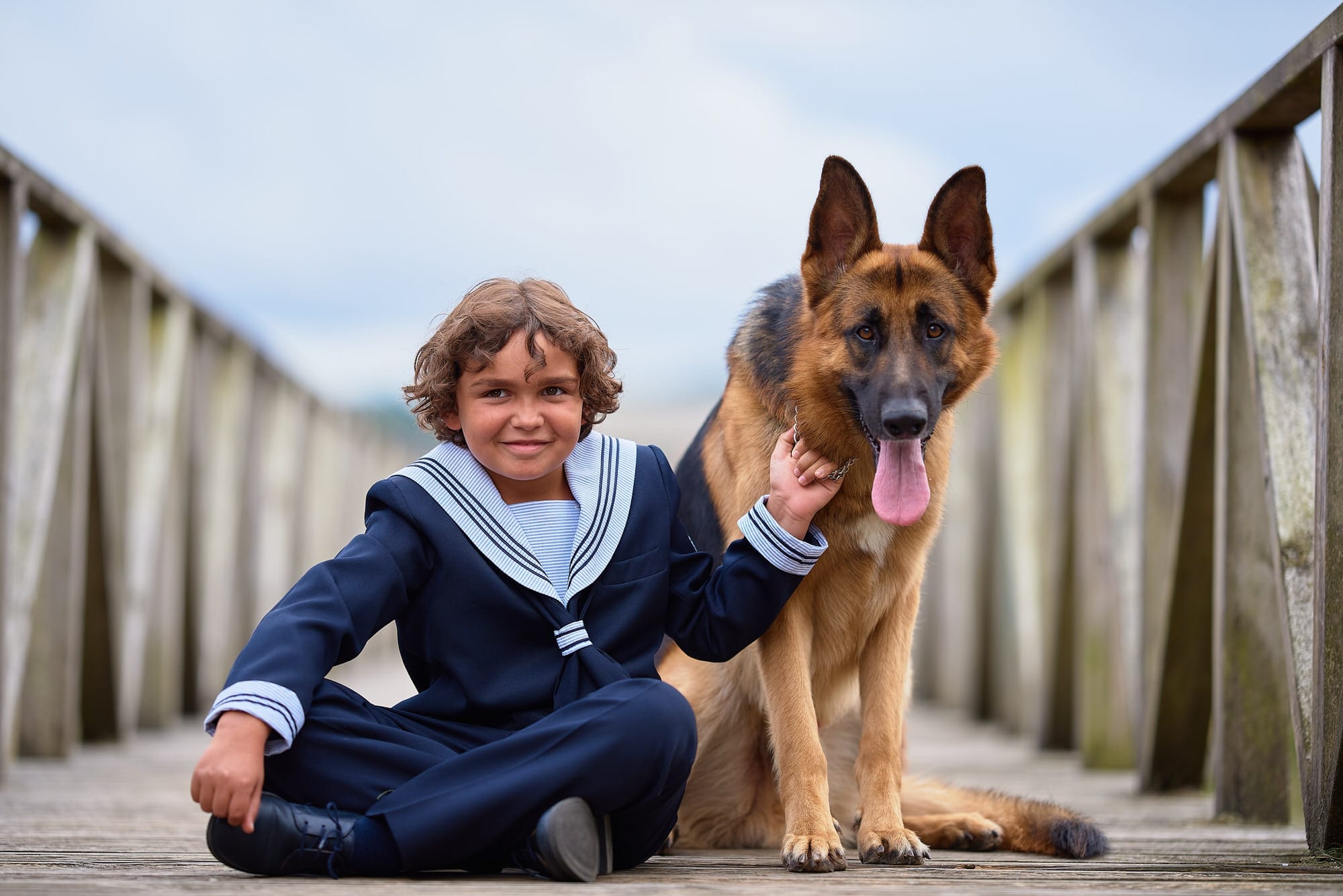 Fotografía de comunión de un niño vestido de marinerito. A su lado tiene a su perro que es un pastor alemán y están sentados en una tarima de madera