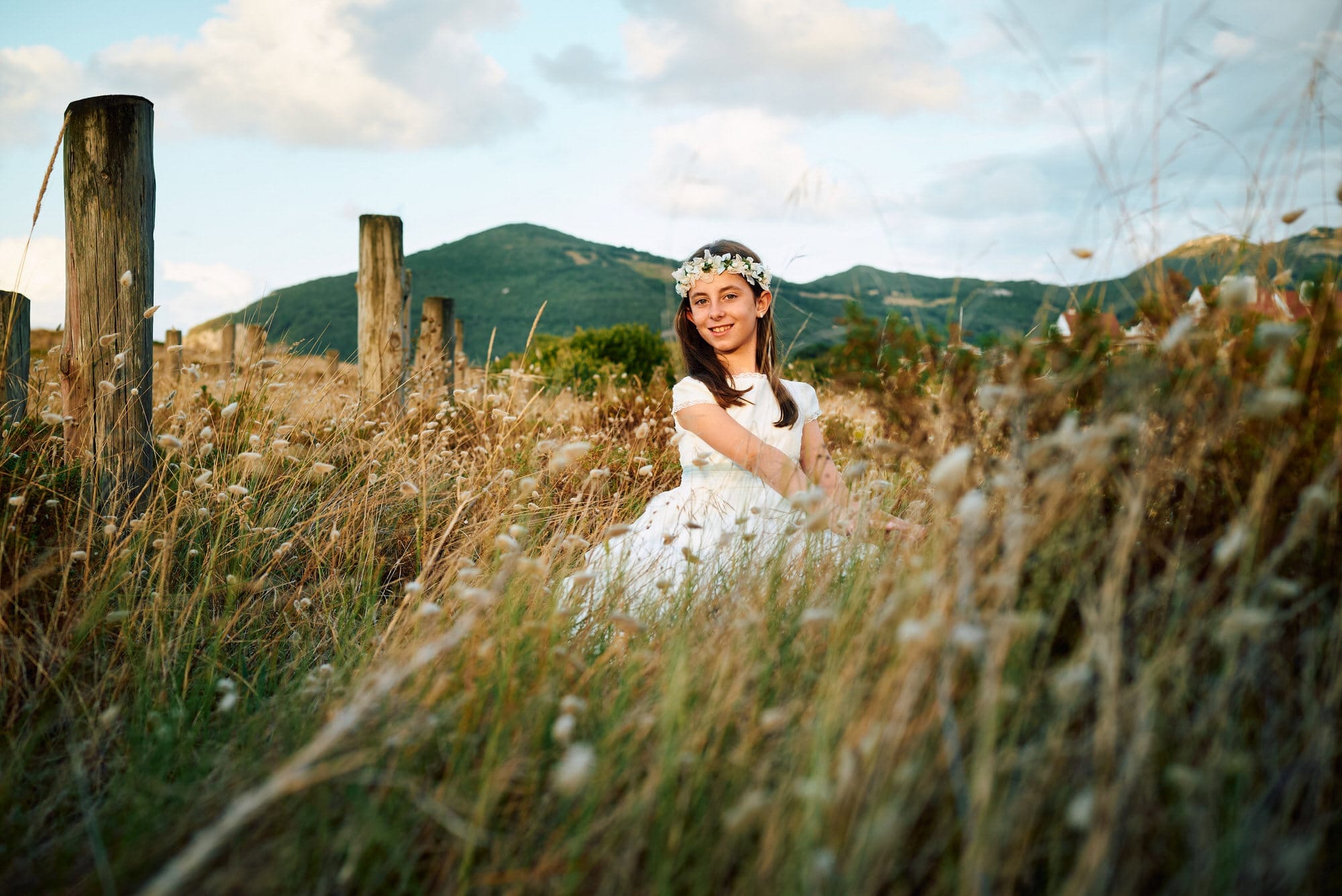 Fotografía de Comunion de una niña en la playa de Berria , ella nos mira sonriendo y está acuclillada entre las hierbas doradas en un espectáculo de luz y color