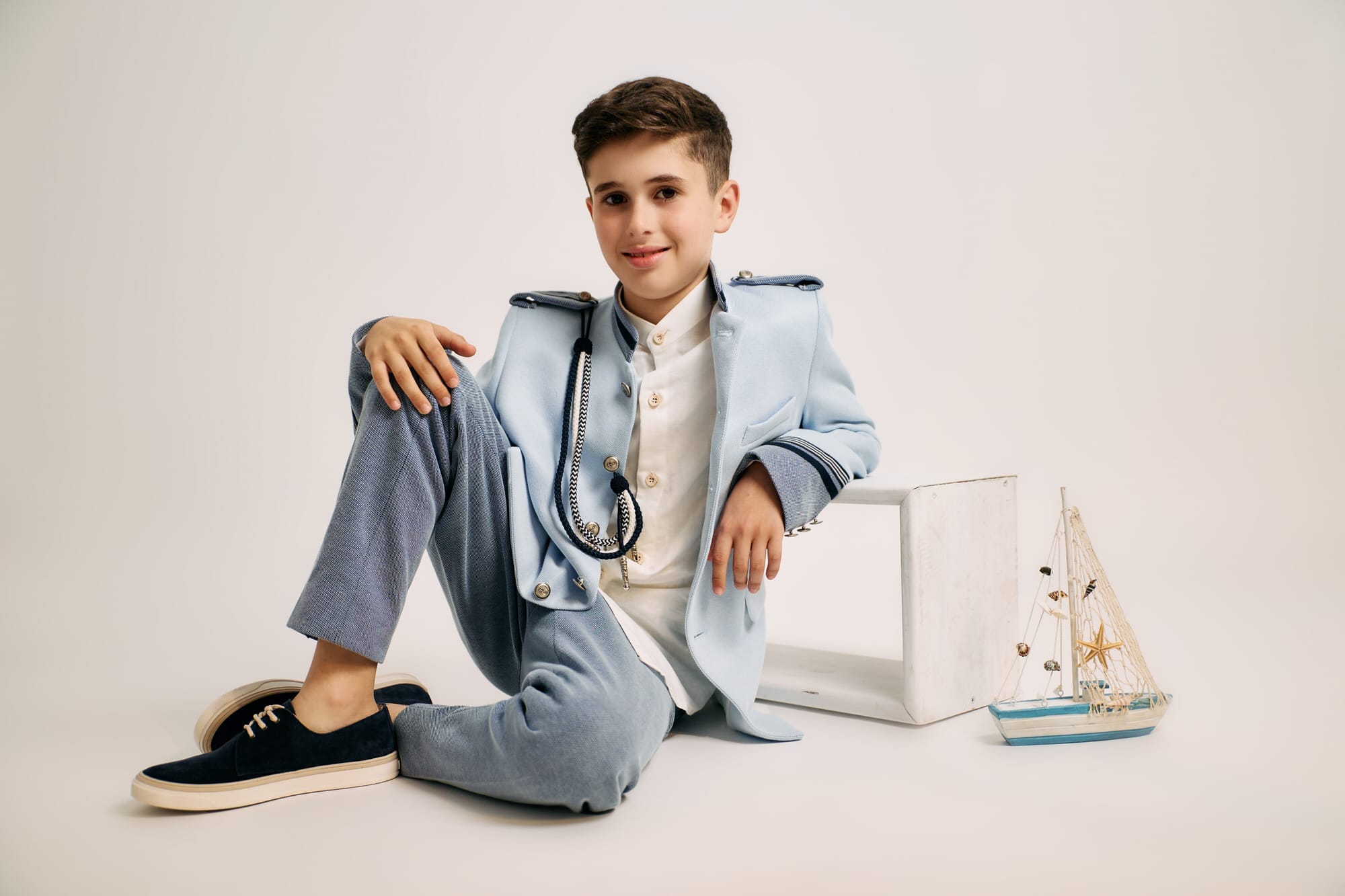 un niño sentado en el suelo del estudio con fondo blanco, lleva un traje de comunion y se ve un barquito velero de juguete