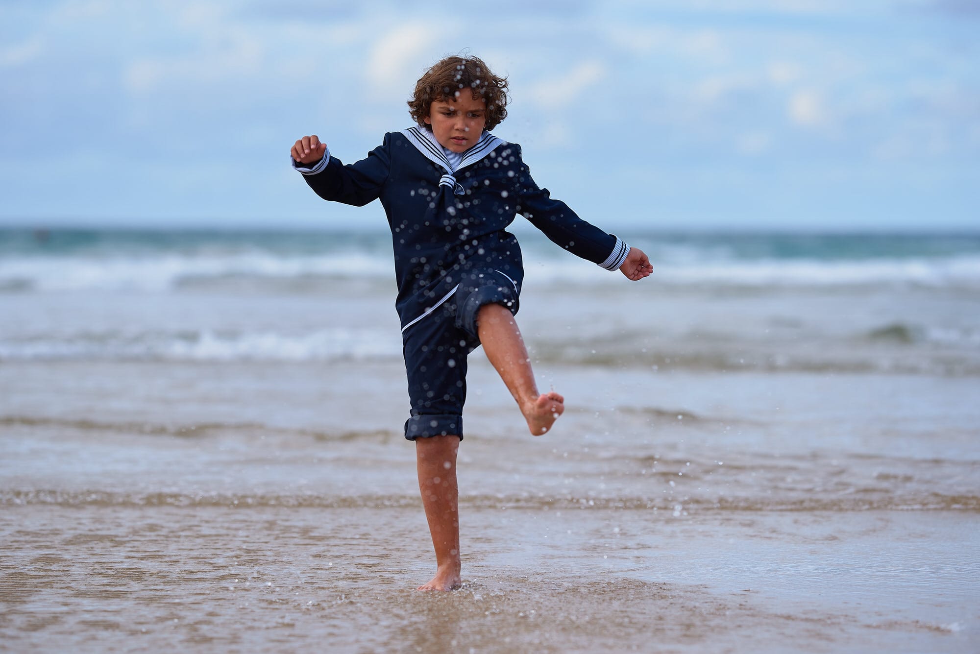 Fotografía de Comunion en la playa de Berria, el niño vestido de marinero salpica con sus pies descalzos, el agua en una actitud dinámica y divertida