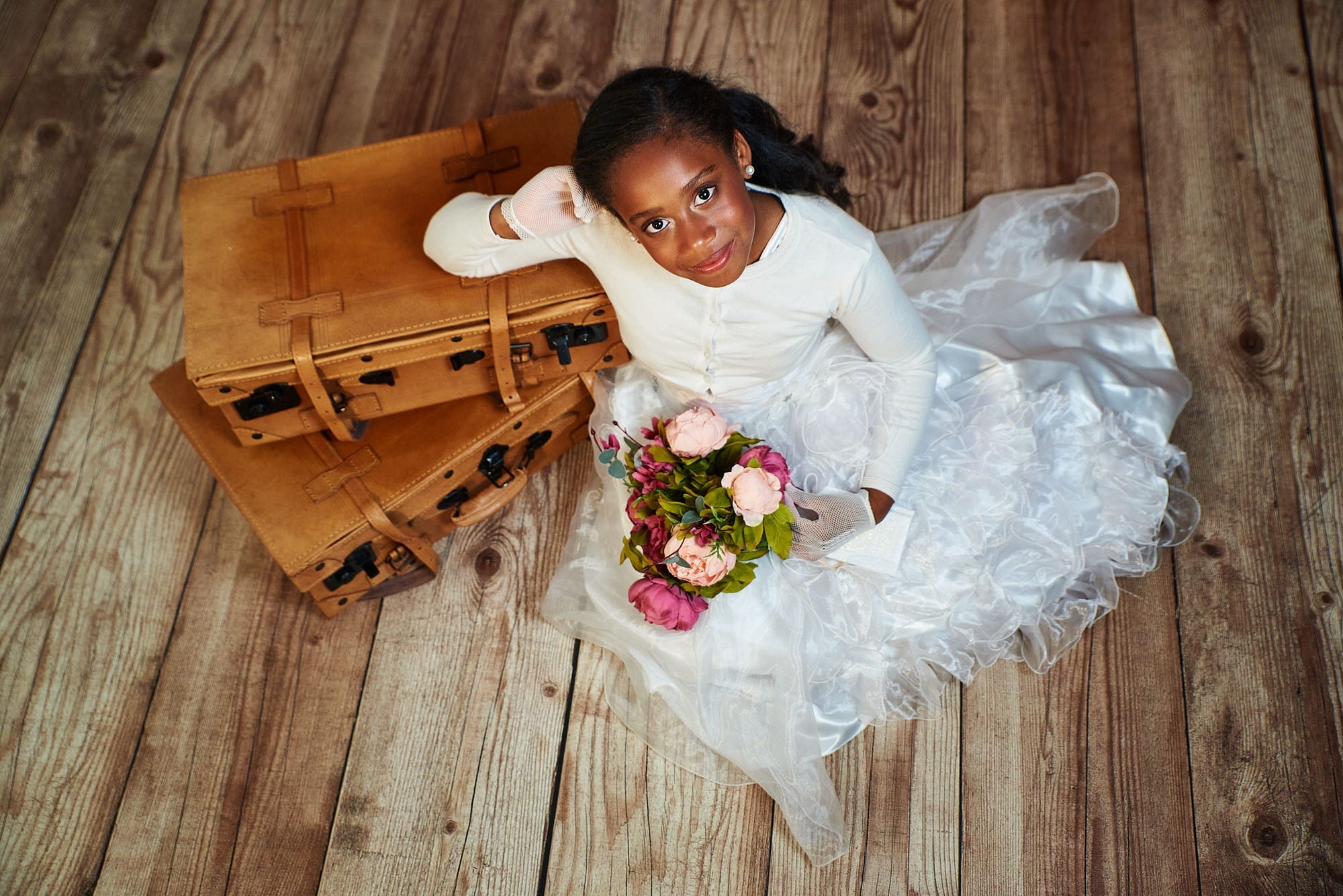 Fotografía de Comunion en Estudio, una preciosa niña, con su blanco vestido sujeta un ramo de Peonías. Sentada en el suelo Se apoya sobre unas maletas de cuero en una foto tomada desde arriba, la niña nos mira mientras sonríe.