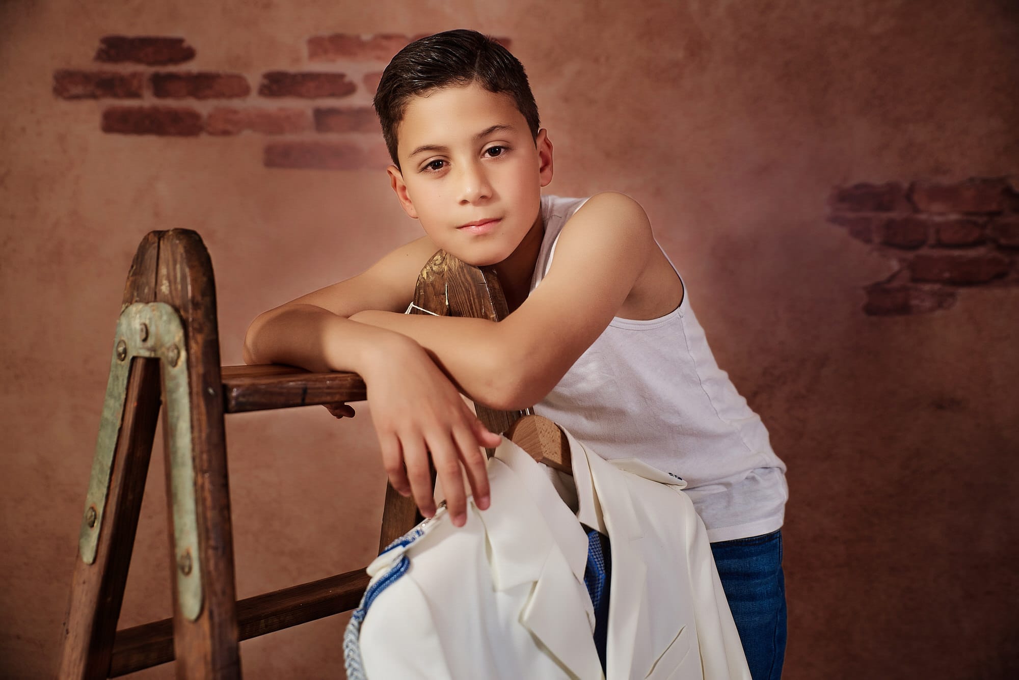 Fotografía de Comunion de Estudio, el niño en camiseta y apoyado sobre una escalera de madera, tipo pintor, nos mira, su traje está colgado de una percha en la escalera