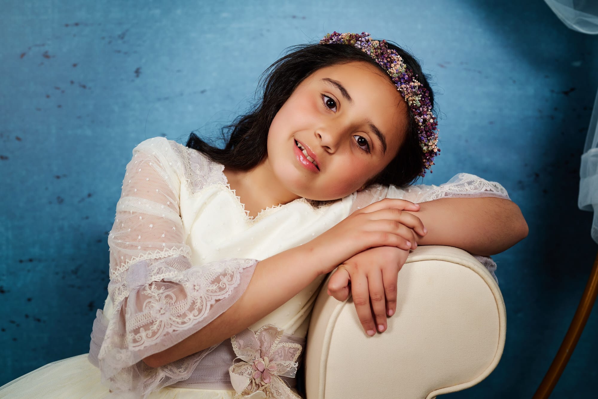 Dulce Fotografía de Comunion, en Estudio , de una niña apoya su cabeza en el brazo de un diván con una mirada dulce contra un fondo azul intenso