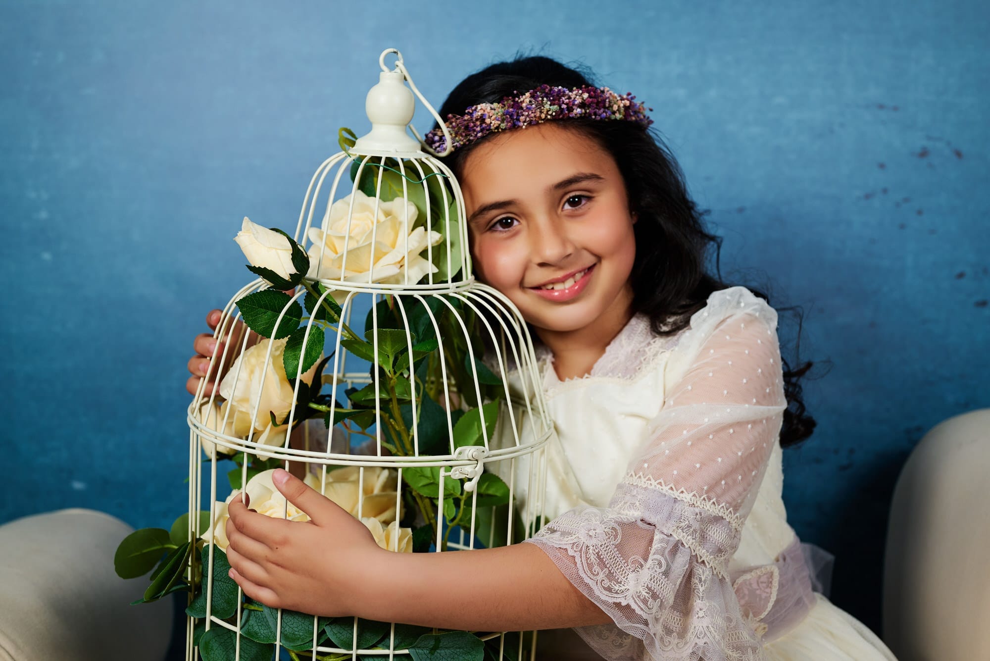 Dulce Fotografía de Comunion, en Estudio , de una niña , abraza una jaula llena de rosas blancas, mientras nos mira dulcemente y nos sonríe para la foto