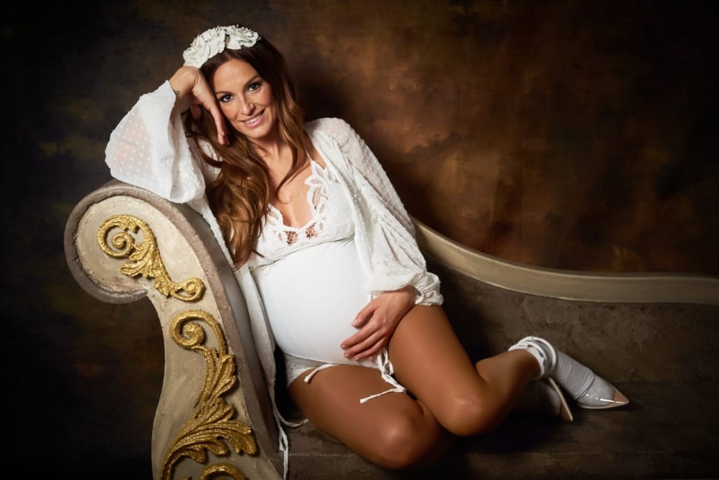 Foto de Embarazo, ella está reposando en un diván luciendo un ramo de Peonías y una diadema a juego en una sesion de estudio que hicimos para ella