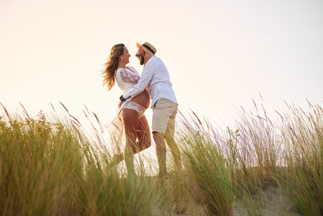 Foto de Embarazo en las dunas de la playa de Laredo ella con un vestido de embarazada y él en pantalón corto y camisa blanca se abrazan , el luce un sombrero de paja tipo canotier