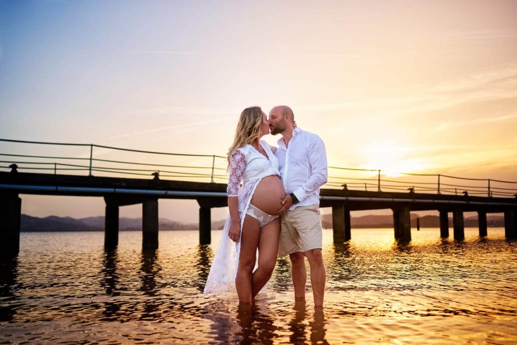 Fotografía de Embarazo al atardecer en el puntal de Laredo, impresionante puesta de sol. Ella luce un vestido de embarazada y el besa a su mujer mientras acaricia su barriga desnuda.