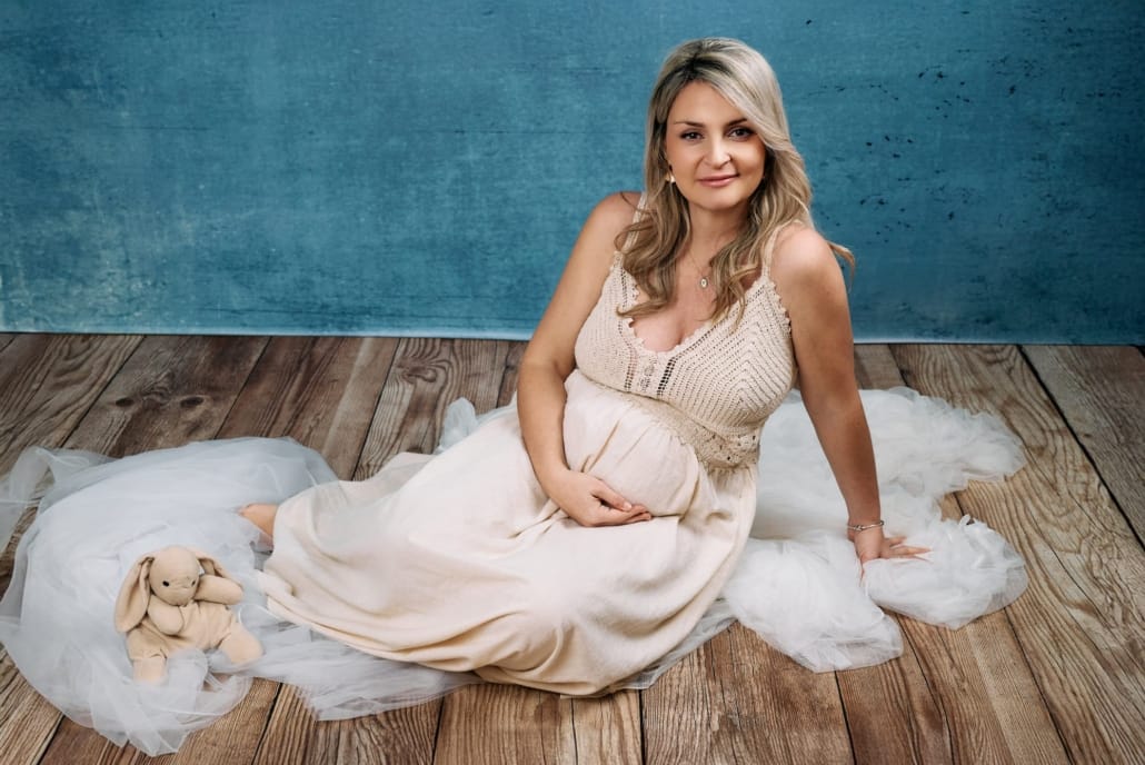 Fotografía de Embarazo en Estudio la novia posa sobre el suelo de madera y un fondo azul con un vestido beis, y tiene una actitud dulce mientras acaricia la barriga