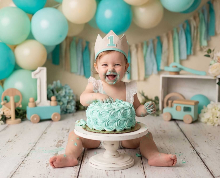 Smash Cake n fotografia de smash cake con ambiente de globos verdes y dorados, es una niña de un año con su tarta , la esta destrozando y comiendo, su carita esta manchada del pastel, el atrezo y el vestuario va a juego con el escenario, lleva una coronita